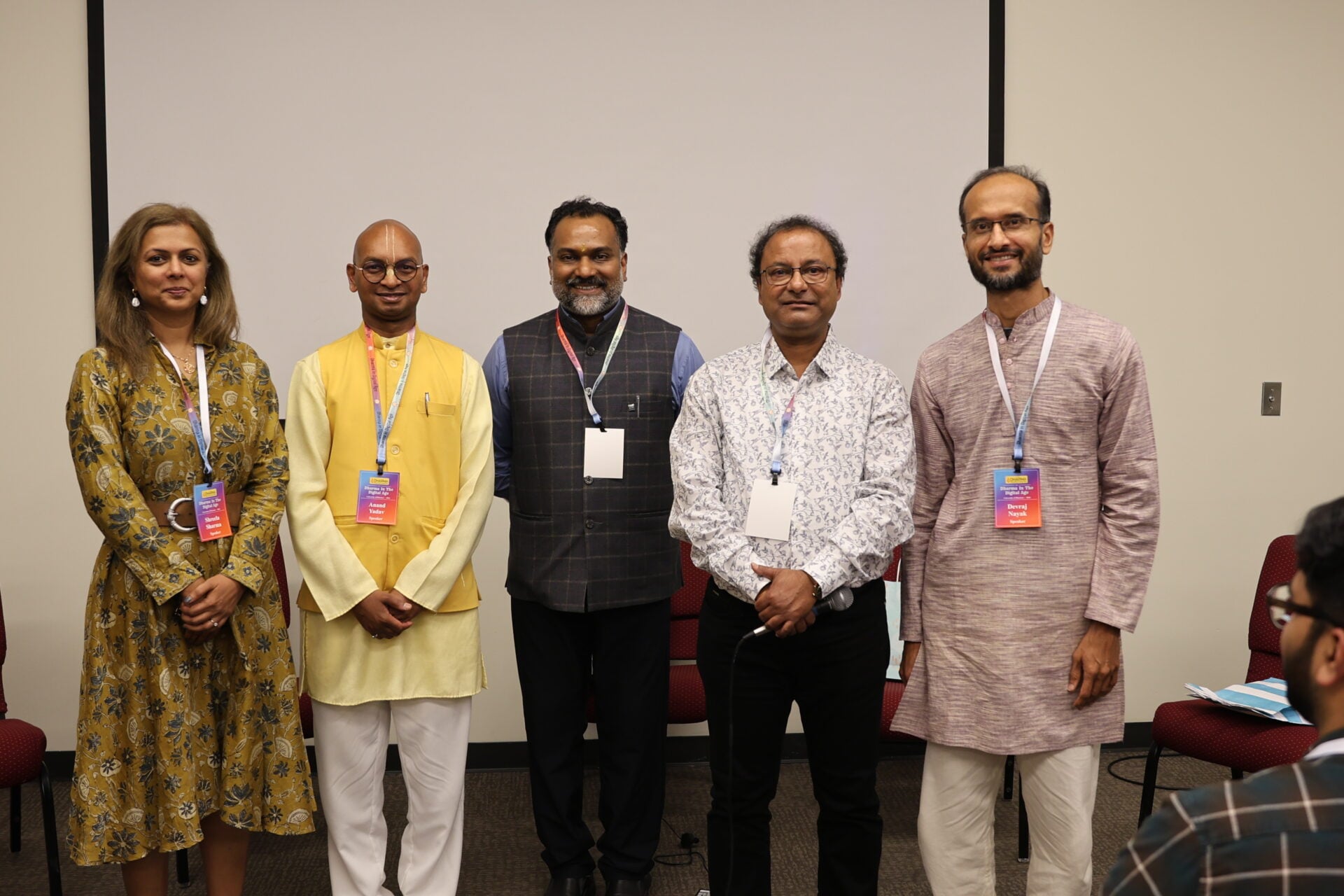 Five Bhagavad Gita event speakers stand together in a conference room, wearing name badges and traditional Indian attire for a health beyond medicine discussion.