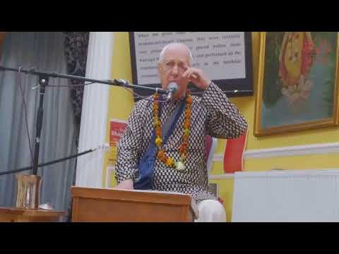 HG Vaiyasaki dasa speaks into a microphone from a wooden lectern during a Srimad Bhagavatam class on SB 8.18.13 in a devotional hall.