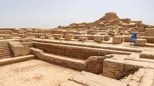 Ancient brick ruins at Mohenjo-daro in Sindh, Pakistan, showing Harappan Indus Valley structures and a mound under a pale desert sky.
