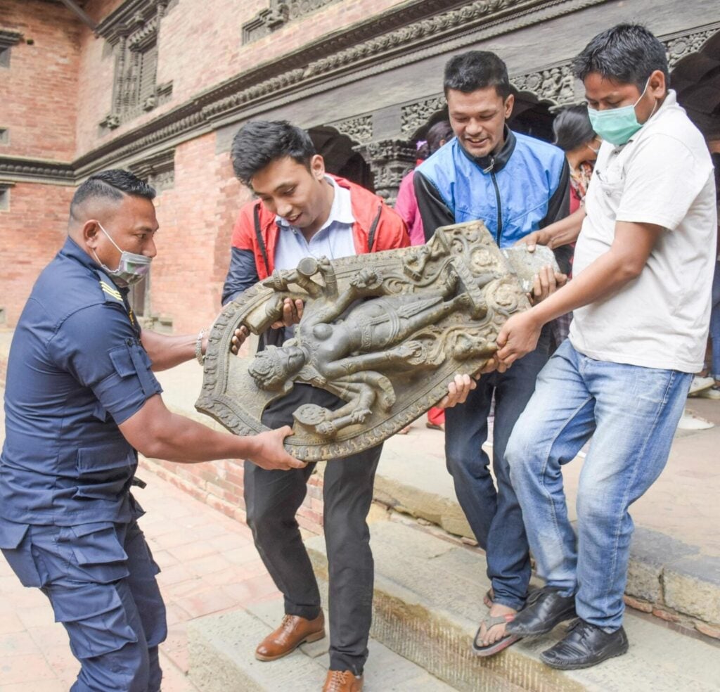 Men carry a carved Hindu deity relief in a Kathmandu heritage courtyard, reflecting Nepal’s sacred art, Hindu Dharma, Bharat ties, and World heritage.