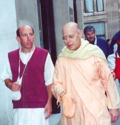 Tamal Krishna Goswami in saffron devotional robes walks outdoors beside a companion in white and maroon clothing, in a memorial photo.