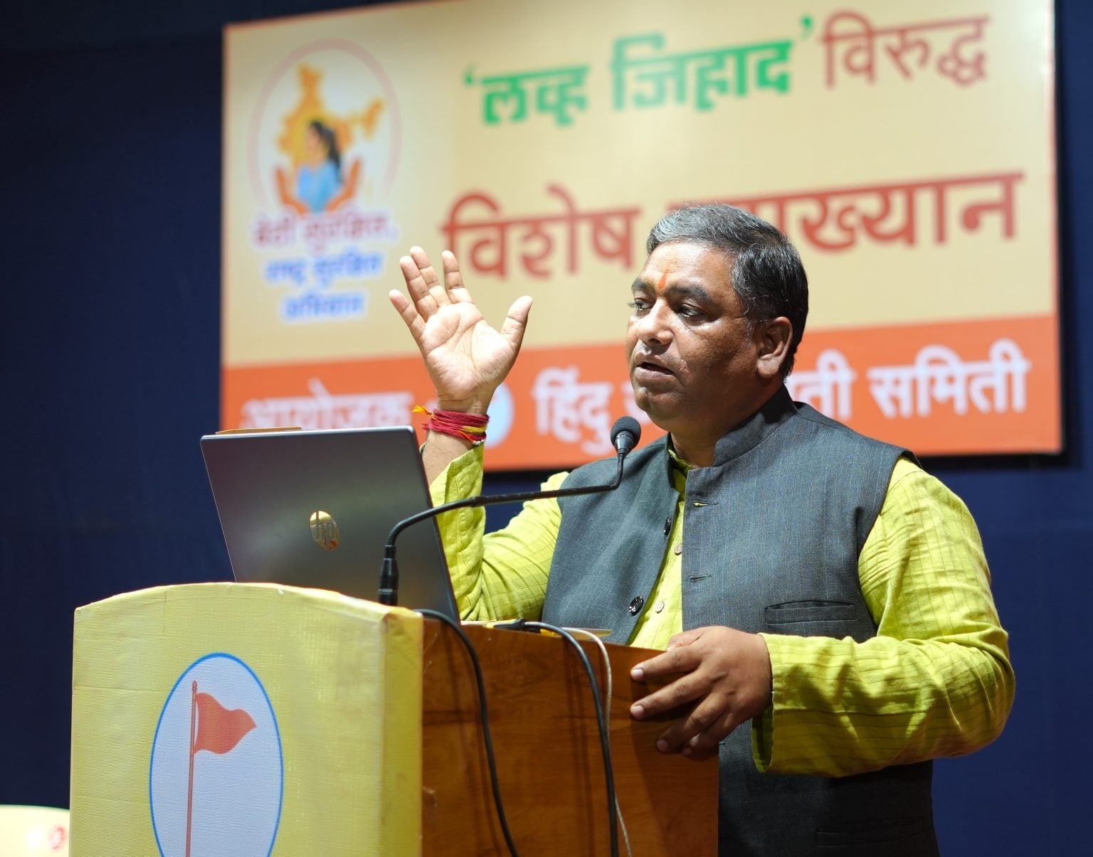 Speaker addresses attendees from a podium during an HJS love jihad awareness lecture in Ichalkaranji, with a campaign banner behind him.