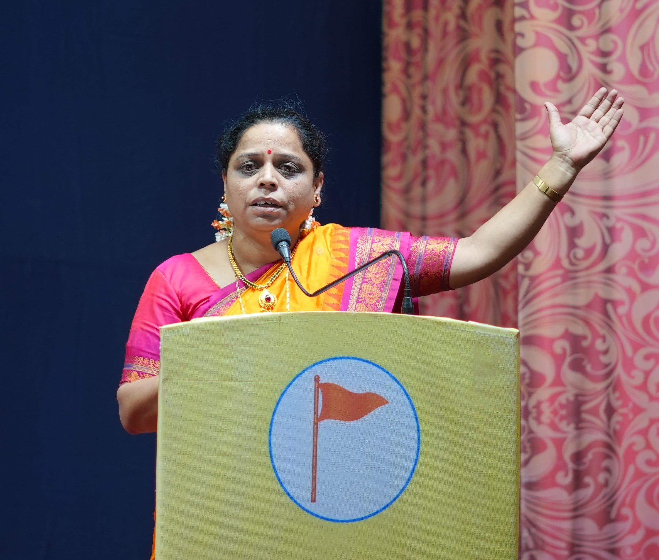 A woman in a bright sari speaks at a podium with a raised hand during a Hindu Jagruti Samiti awareness lecture in Ichalkaranji.