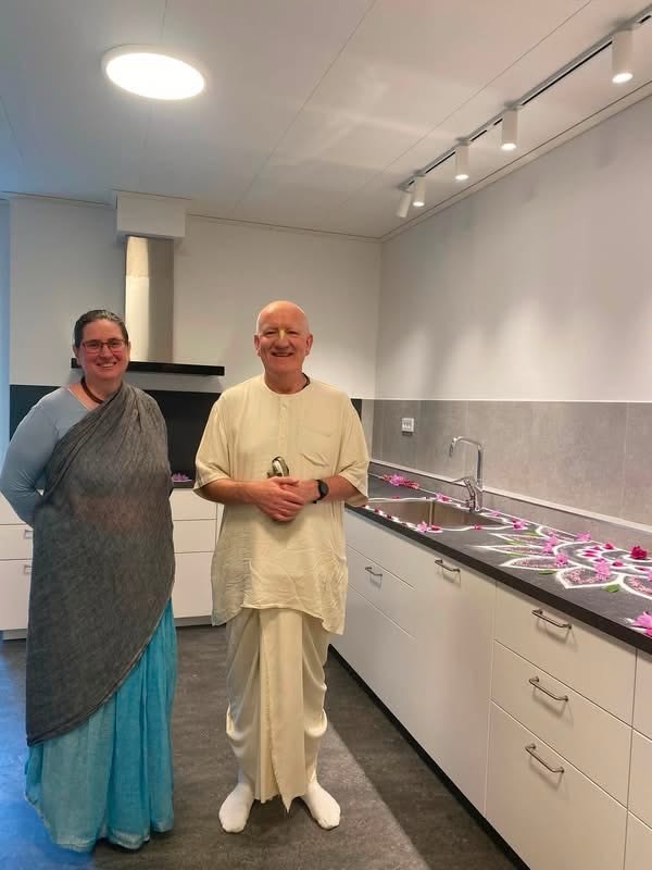 Two devotees stand in a newly renovated ISKCON Almvik temple kitchen with white cabinets, sinks, track lighting, and flower petals on the counters.