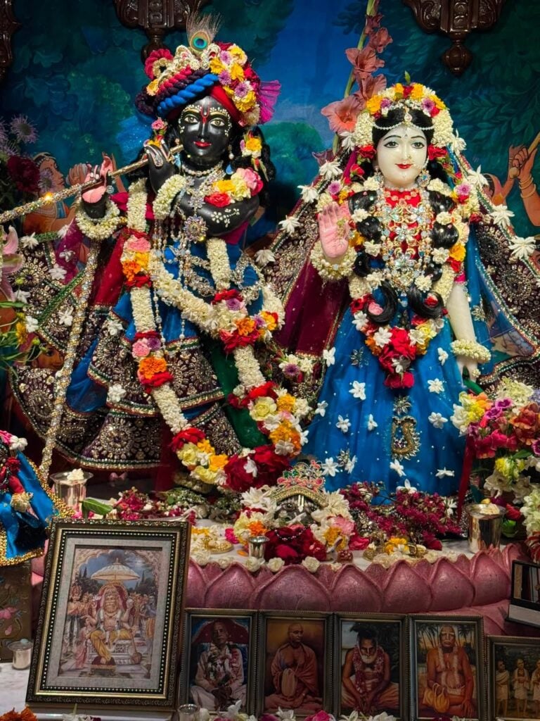 Decorated Radha Krishna deities on a temple altar with flower garlands, ornate clothing, framed devotional portraits, and offerings