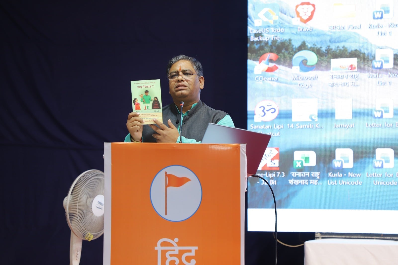 A speaker at a Hindu Jagruti Samiti event in Belagavi holds a booklet at an orange podium during a lecture on community awareness.