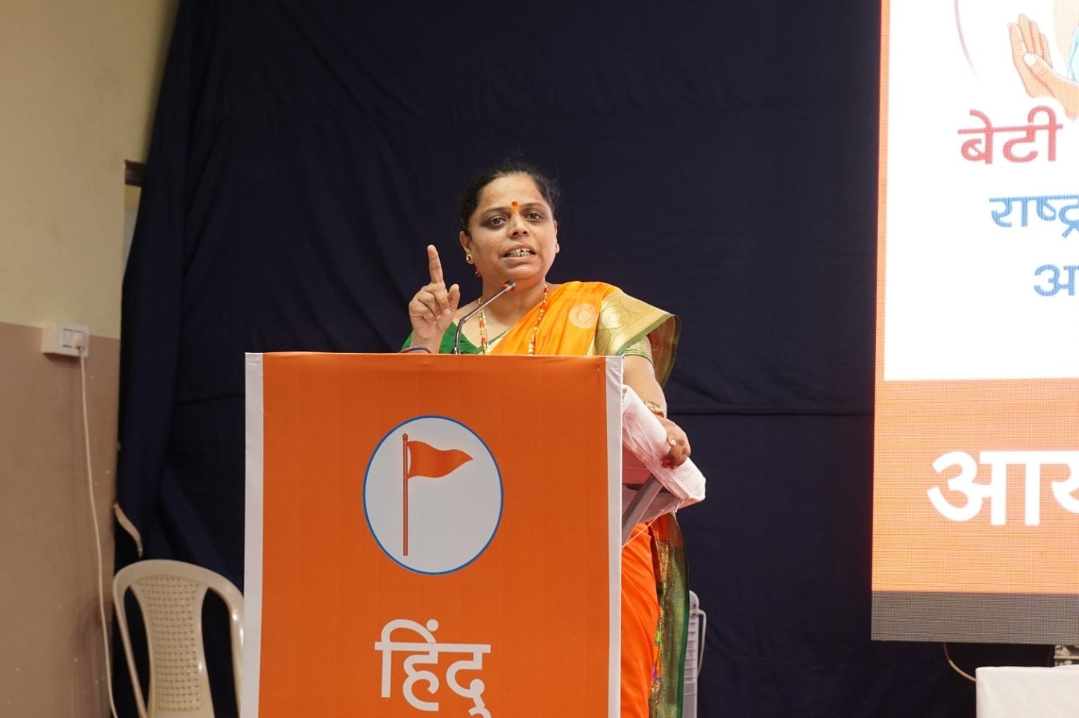 A woman speaker in a saffron sari addresses a Belagavi Hindu Jagruti Samiti gathering from an orange podium on love jihad awareness.