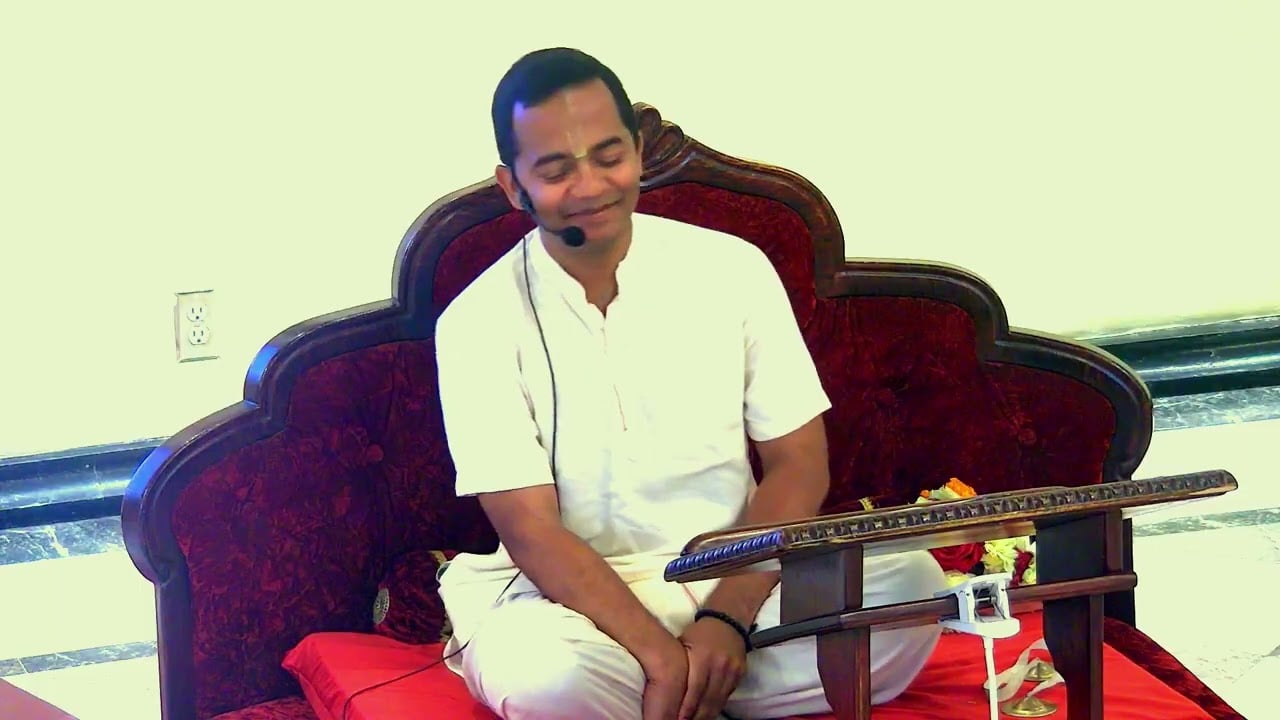 Shyamcandra Prabhu seated in white attire with a headset microphone during SB 4.8.65 class at ISKCON Los Angeles.