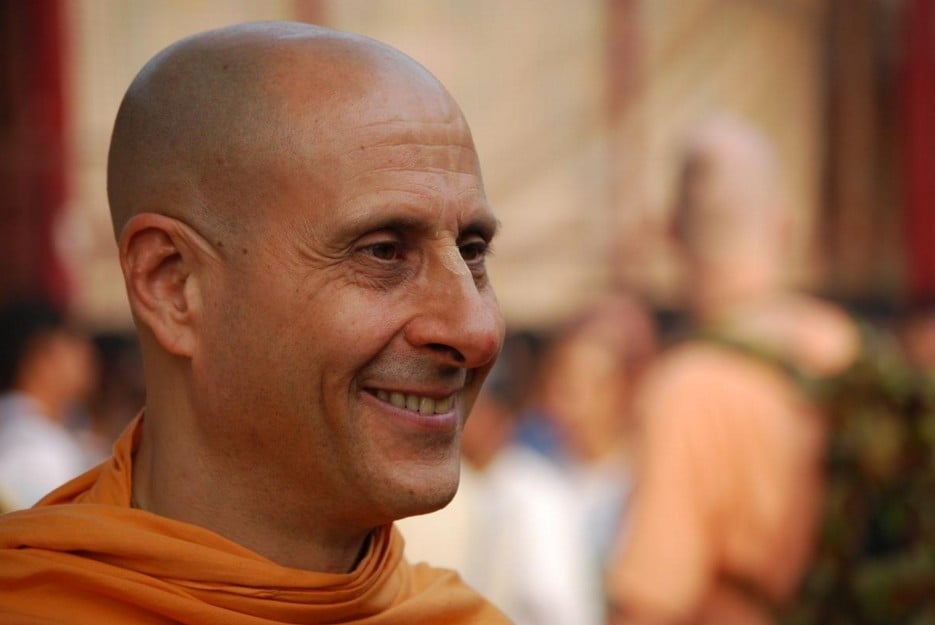 Radhanath Swami smiles in saffron robes, shown in close-up against a softly blurred gathering of devotees in warm orange tones.