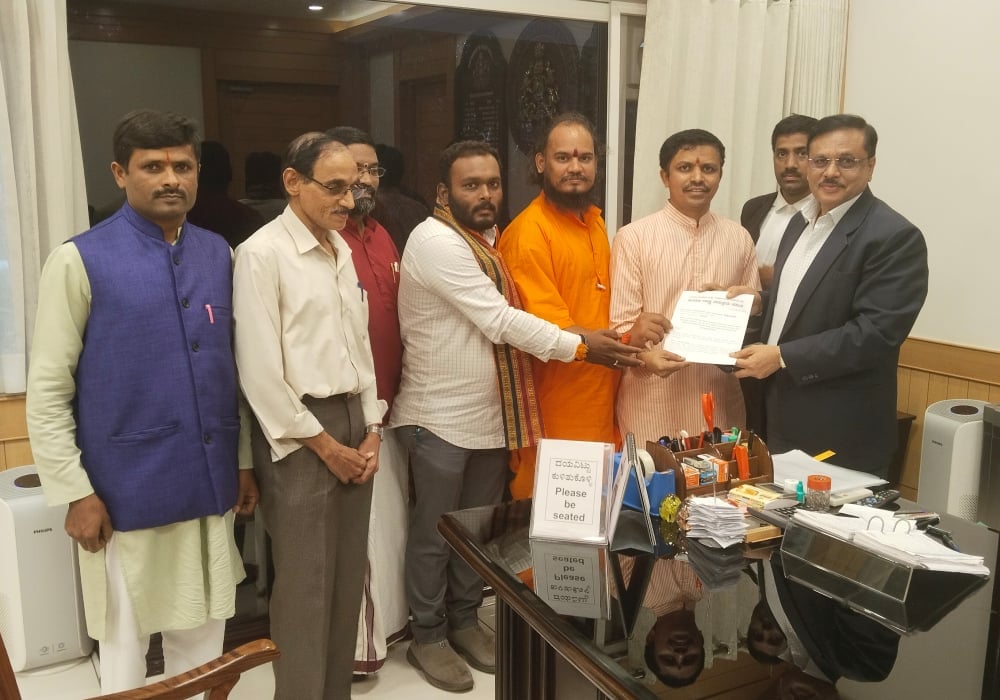 Eight men stand in an office as representatives present a memorandum to a suited official concerning legal action over denigration of Hindu deities.