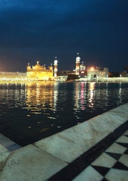 The illuminated Golden Temple complex in Amritsar reflected in the sacred pool at night, viewed from a marble walkway along the water.
