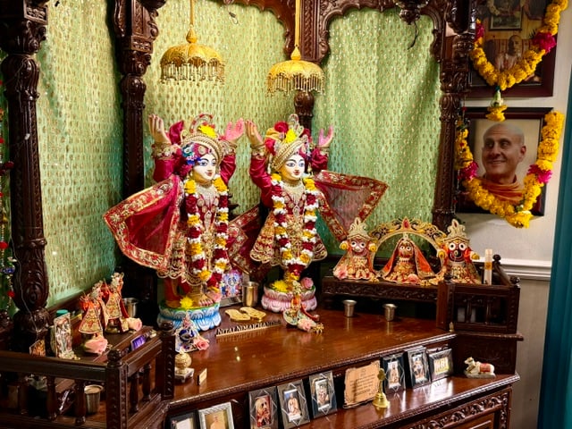 Sri Sri Nitai Nimai Chandra deities in ornate red garments and flower garlands, standing with raised arms on a carved wooden home altar in Seattle.