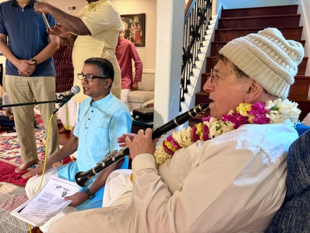 Ramai Swami, wearing a flower garland and knit cap, plays clarinet beside a seated singer during the Nitai Nimai Chandra anniversary bhajan.