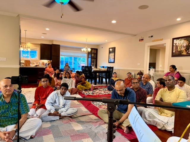 Devotees seated on rugs in a Seattle home during the Sri Sri Nitai Nimai Chandra anniversary gathering, with microphones arranged for talks and bhajans.