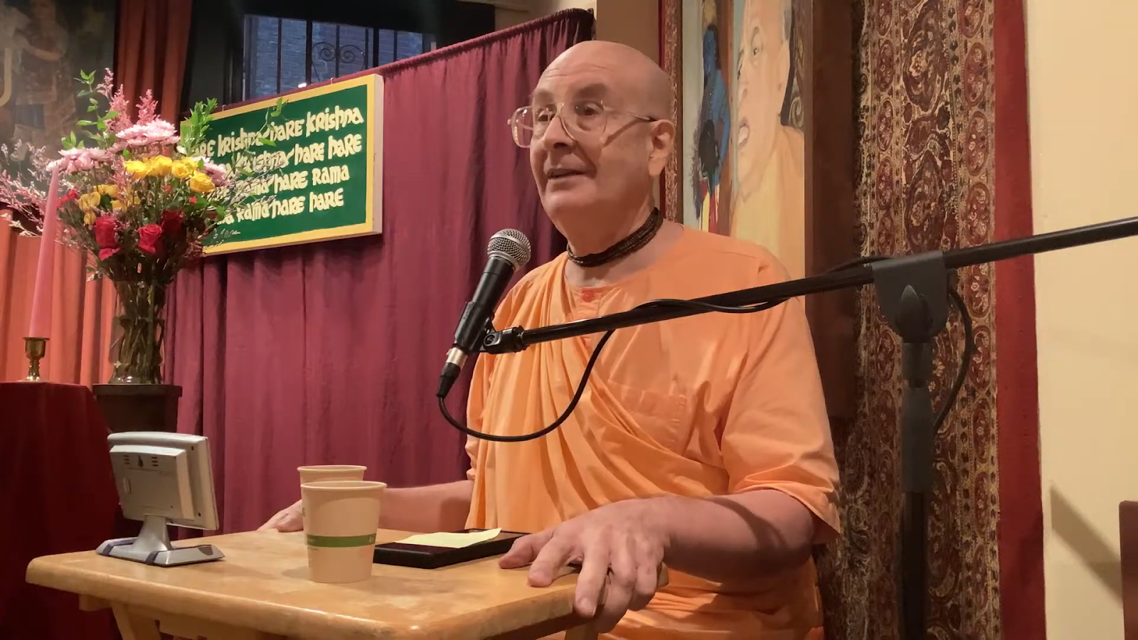 ISKCON monk in saffron robes speaks into a microphone at a temple lectern during Day 1 of ISKCON’s 60th Anniversary Week.