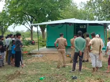 कौशांबी के ग्रामीण इलाके में हरे रंग की अस्थायी मजार के पास पुलिसकर्मियों और स्थानीय लोगों की भीड़, पेड़ों से घिरा घटनास्थल।