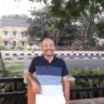 Older man in a navy, white and pale pink polo shirt standing beside a railing outdoors, accompanying a book review on Indic kingship and Marxist historiography.