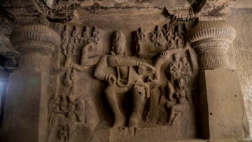 Weathered stone relief of a multi-armed deity and attendants framed by carved pillars inside an ancient Indian rock-cut temple.