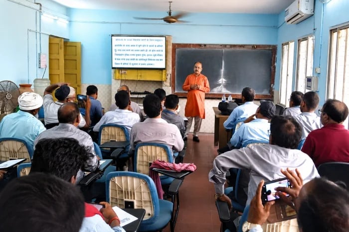 A man in an orange kurta addresses seated attendees in a classroom equipped with blackboards, a wall-mounted screen, windows and an air conditioner.