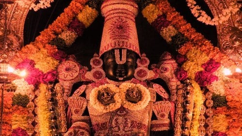 Venkateshwara Swamy idol at Tirumala, richly adorned with a tall crown, sacred forehead marks, gold jewelry, flower garlands and glowing temple lamps.