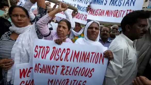 Women lead a crowded protest in Pakistan, holding a placard that reads “Stop discrimination against religious minorities” as demonstrators gather behind them.