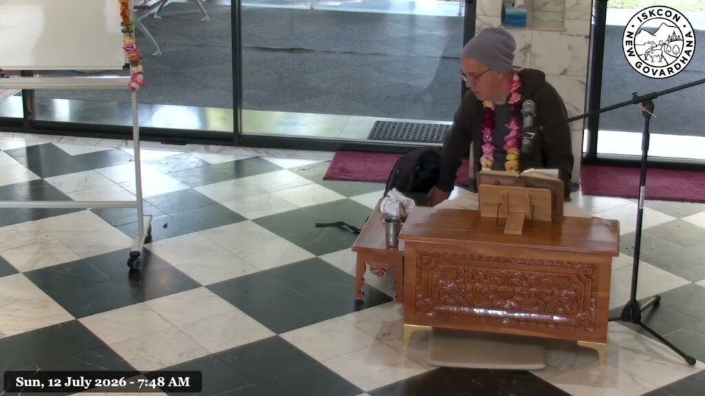Nrsimha Kavaca Das, wearing a flower garland, sits beside a microphone and scripture at a carved wooden lectern in ISKCON New Govardhana Temple.