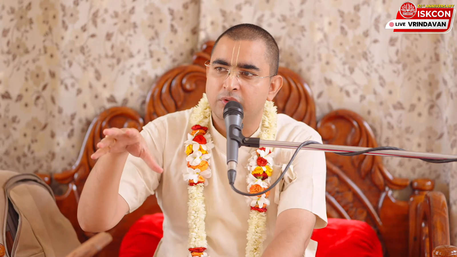 His Grace Rupanuga Bhakta Prabhu, wearing Vaishnava tilak and a flower garland, speaks into a microphone during a Caitanya-caritāmṛta Ādi-līlā class.