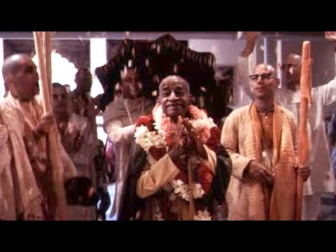 Srila Prabhupada, wearing flower garlands and holding his palms together, walks among saffron-robed devotees at a Hare Krishna gathering.