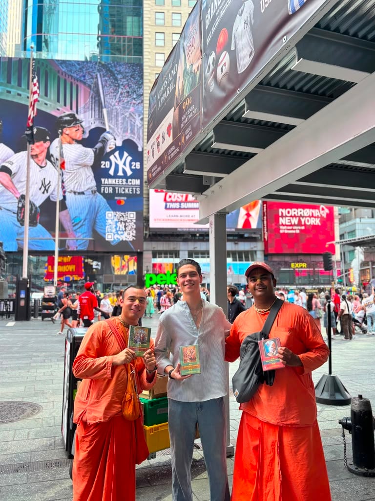 Two Krishna devotees in orange robes pose with a smiling young man holding spiritual books amid the crowds and billboards of Times Square.