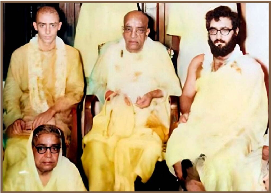 Vintage photograph of Srila Prabhupada seated in pale saffron robes with several attendees, including a bearded gentleman at his right.