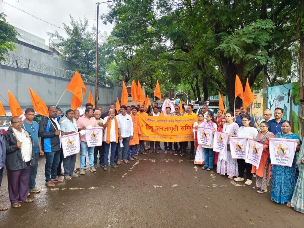 Protesters in Ichalkaranji gather on a wet, tree-lined street with saffron flags, placards and a Hindu Janajagruti Samiti banner opposing “love jihad.”