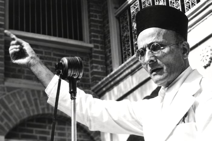 Black-and-white photograph of Swatantryaveer Vinayak Damodar Savarkar in a black cap and round glasses, pointing while speaking at a vintage microphone.
