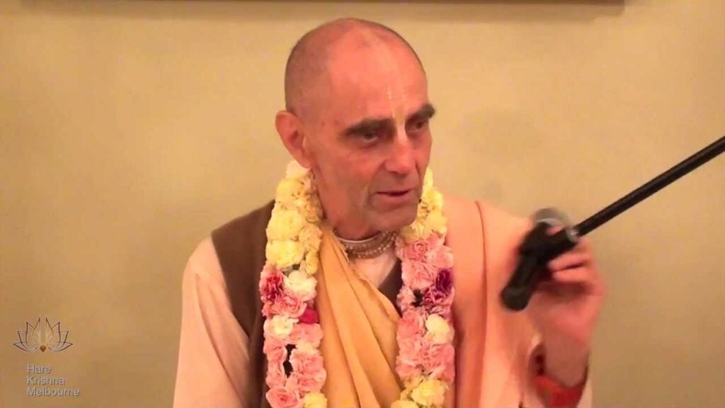 A saffron-robed Hare Krishna monk wearing a flower garland and tilak holds a microphone while speaking, with a Hare Krishna Melbourne logo visible.