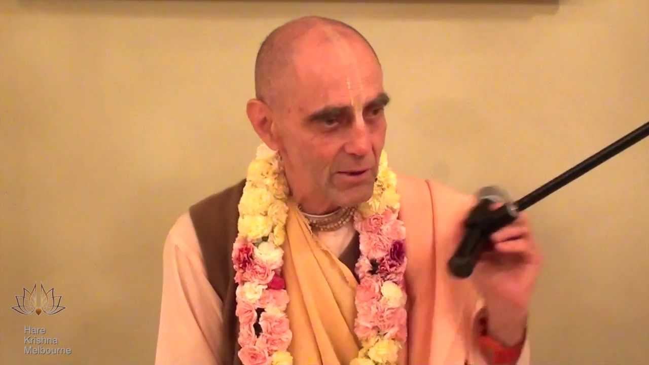 A saffron-robed Hare Krishna monk wearing a flower garland and tilak holds a microphone while speaking, with a Hare Krishna Melbourne logo visible.