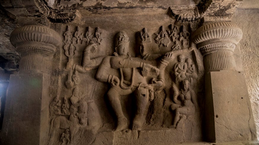 Weathered stone relief of a multi-armed Hindu deity, surrounded by attendants and framed by ornate pillars inside an ancient temple in Bharat.