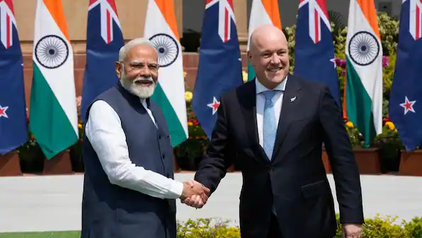 Prime Minister Narendra Modi and New Zealand Prime Minister Christopher Luxon shake hands before alternating Indian and New Zealand flags.