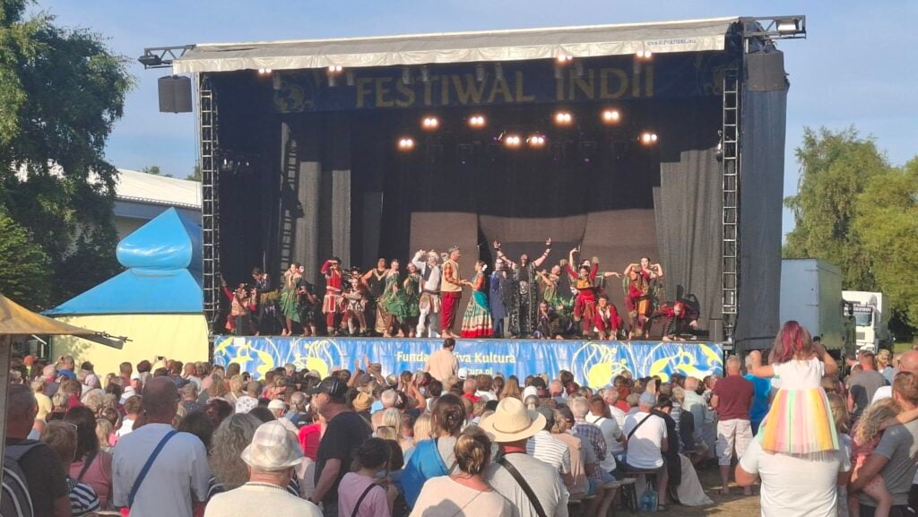 A large crowd watches costumed performers pose on an outdoor stage beneath a Festival Indii banner at the Hare Krishna India Festival in Poland.