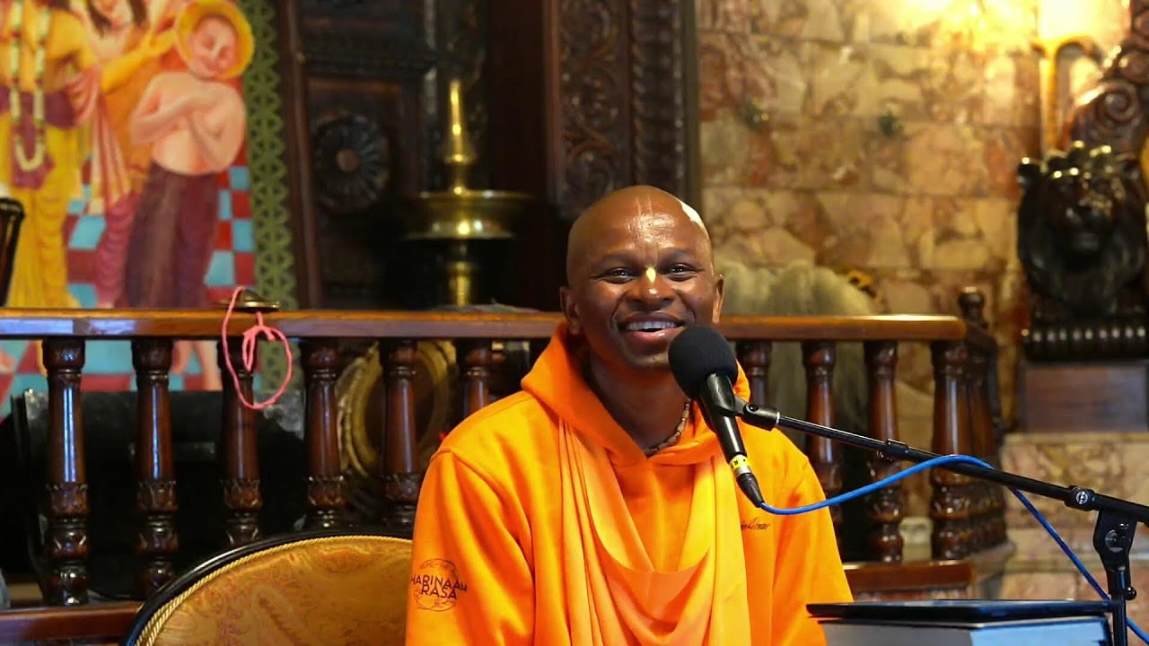 HH Bhakti Sarvajna Gauranga Swami smiles while speaking into a microphone in saffron robes during a Srimad Bhagavatam 4.24.49 lecture.
