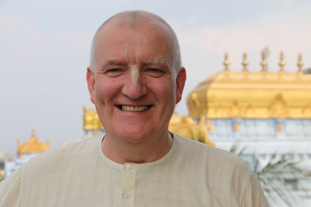Portrait of Praghosha Das smiling in cream devotional attire with Vaishnava tilaka, standing before a softly focused golden-domed temple.