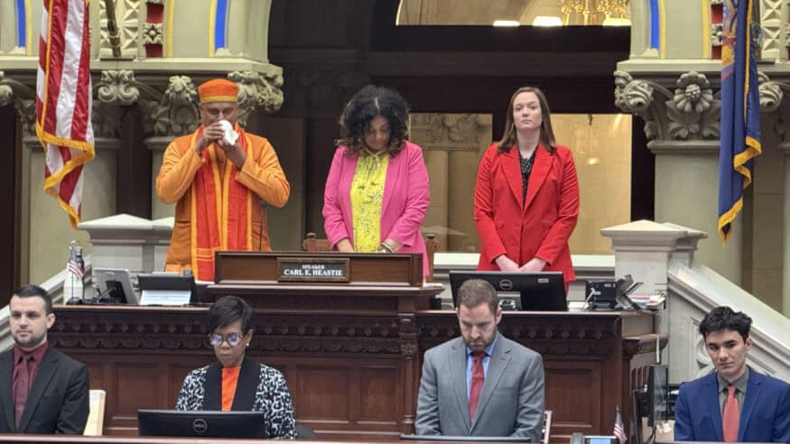 Yogi Dileep, dressed in saffron robes, blows a conch in the New York State Assembly chamber as officials stand behind and legislators sit below.