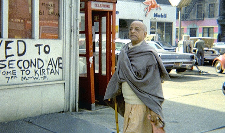 Srila Prabhupada, wrapped in a gray shawl and holding a cane, walks along a New York street beside a red telephone booth and parked cars.