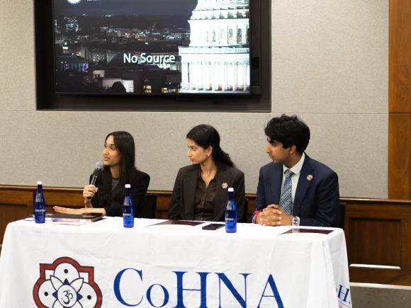 Three adults sit at a CoHNA-draped table as a woman speaks into a handheld microphone.