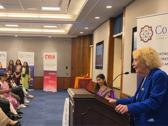Elderly woman in a blue jacket speaks at a lectern to a seated audience in a conference room.