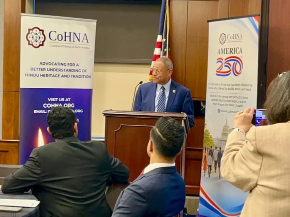 A man speaks at a lectern between CoHNA and America 250 banners as seated attendees listen.