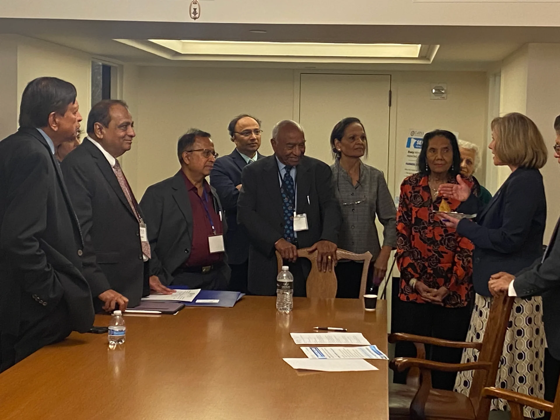 Adults gather around a conference table as a woman on the right speaks and gestures.