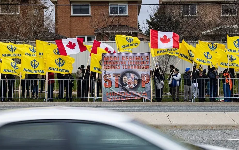 Crowd behind a roadside railing holding yellow Khalistan flags, Canadian flags and a protest banner