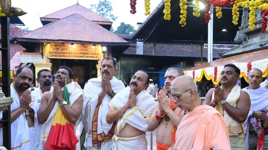 Men in white traditional garments pray with folded hands beside a saffron-robed elder in a decorated courtyard.