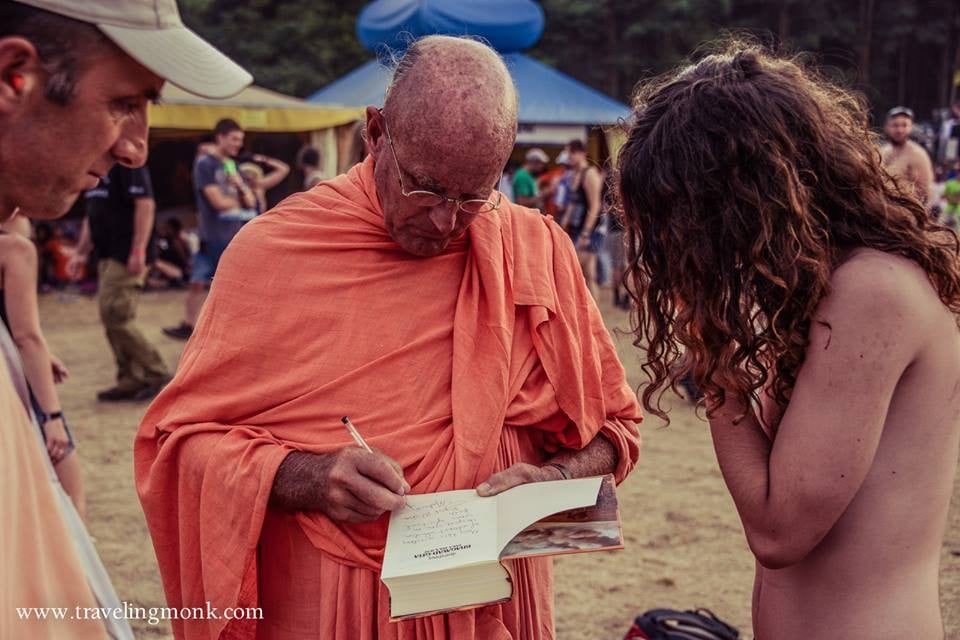 Elderly man in orange robes signs an open book as two people watch at an outdoor gathering.