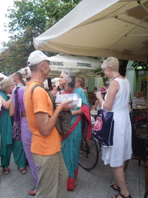 Man in an orange shirt holding books amid a crowd beneath large outdoor cafe umbrellas.