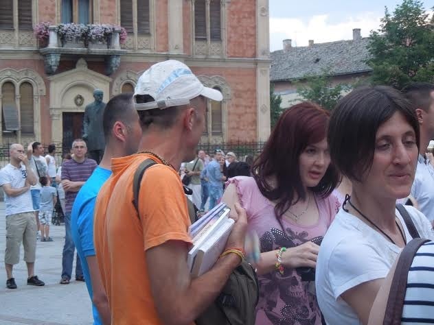 Man in an orange shirt and white cap holds books among a crowd outside an ornate building.
